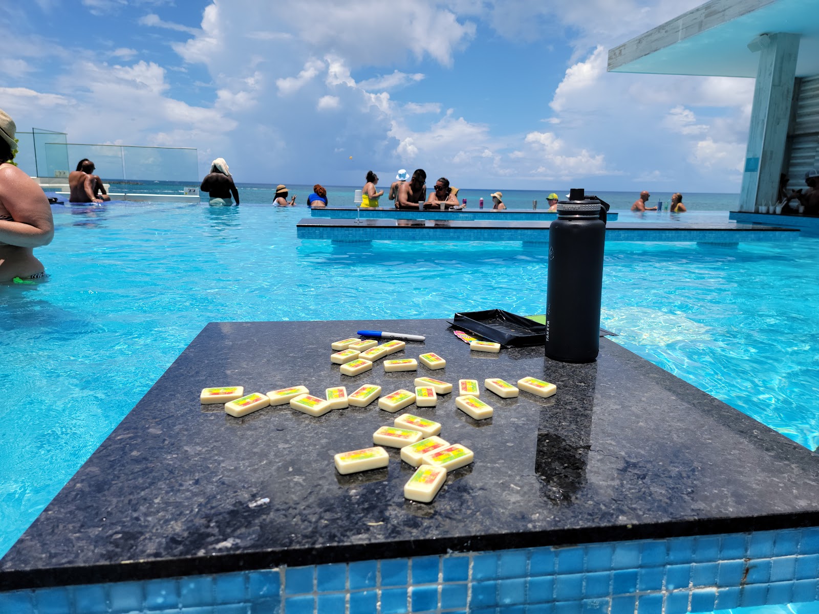 Swim lesson platform at Hotel Riu Palace Paradise Island with kickboards arranged on a dark table, turquoise pool, ocean, and guests in background.