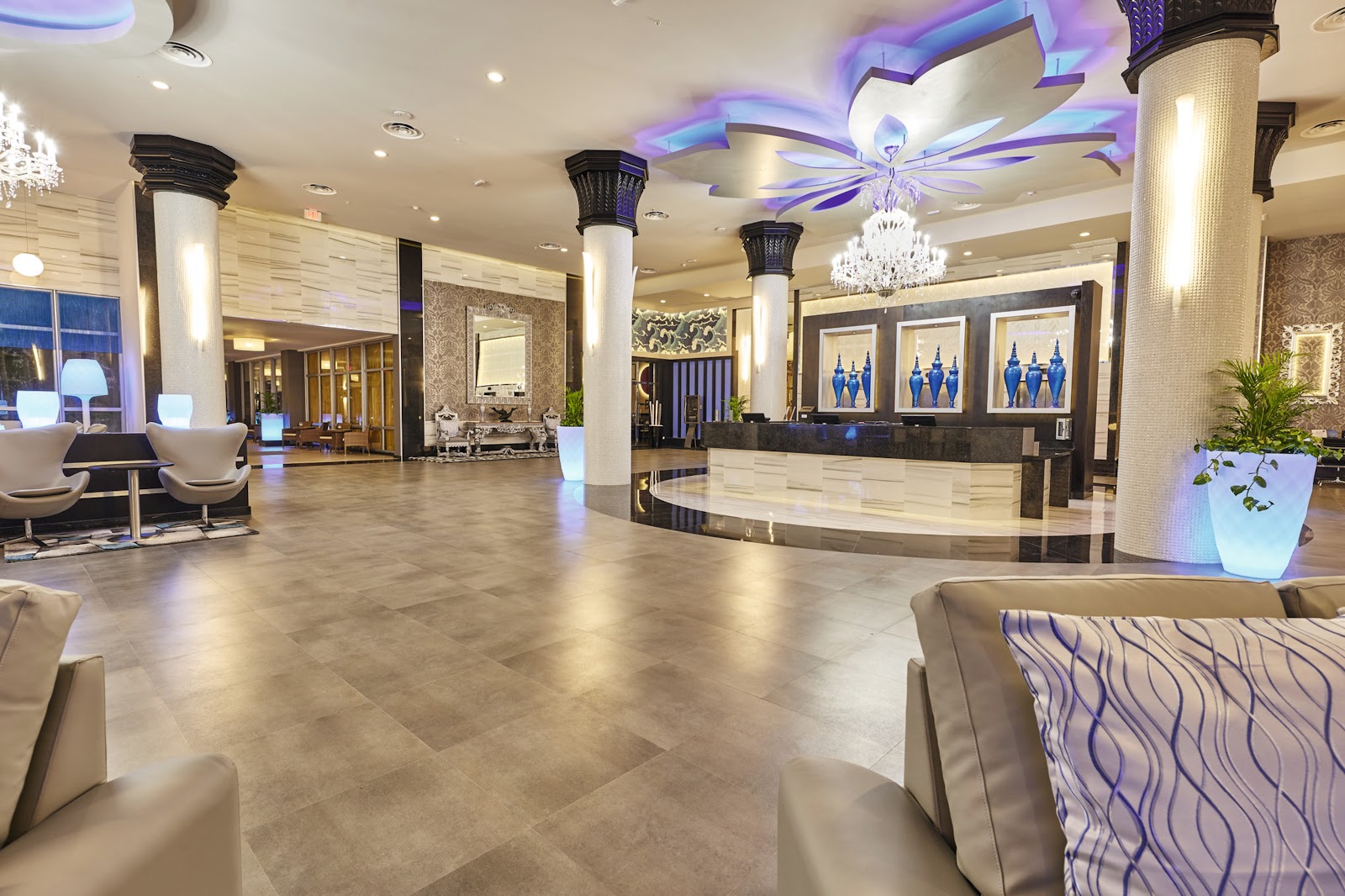 Spacious resort lobby with polished tile flooring, grand chandelier, columned archways, purple-lit geometric ceiling details, and reception desk with blue artwork.