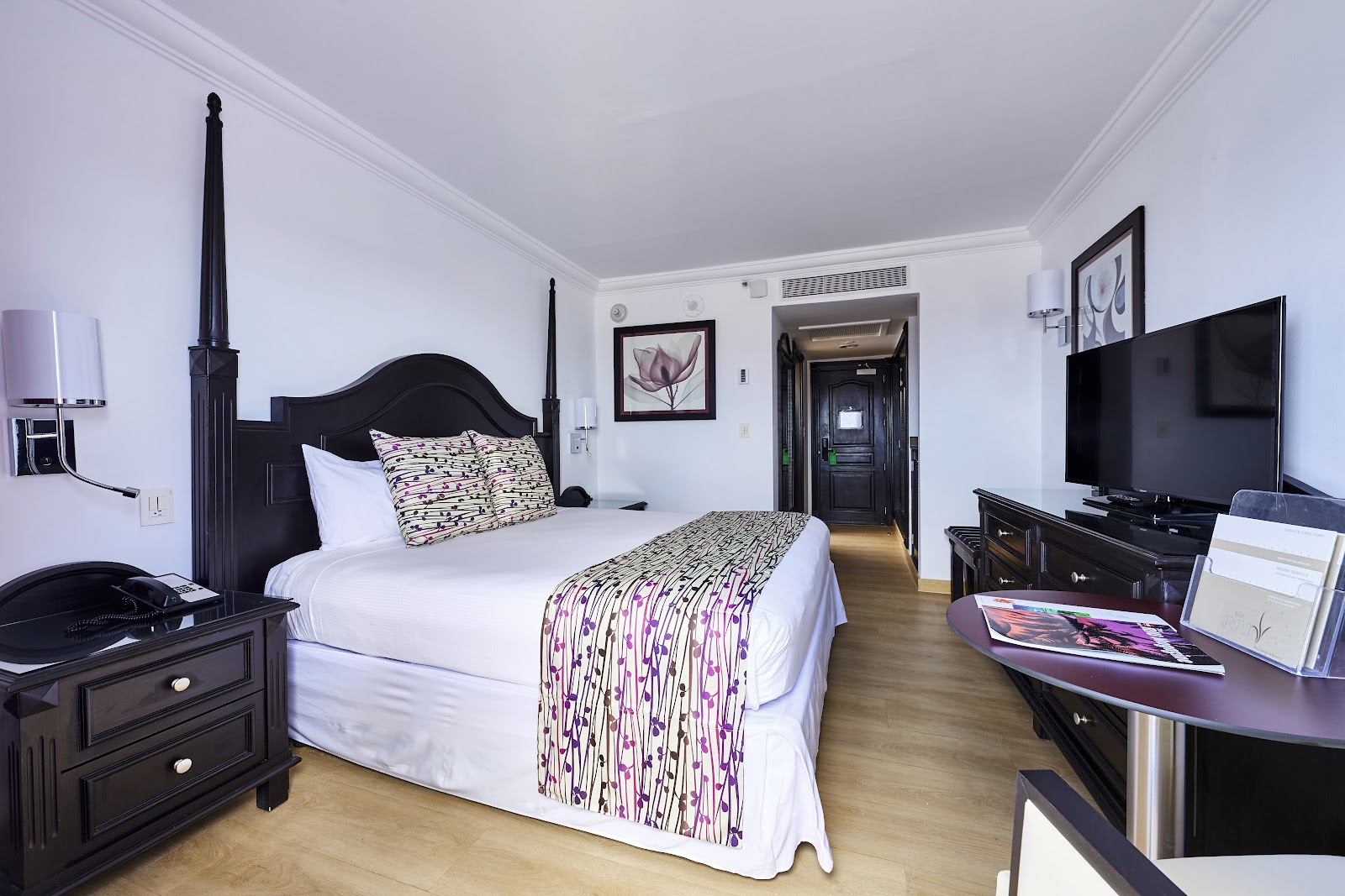 Hotel room with black four-poster bed, white linens, patterned purple bedcover, flat-screen TV, black furniture, and wood flooring.