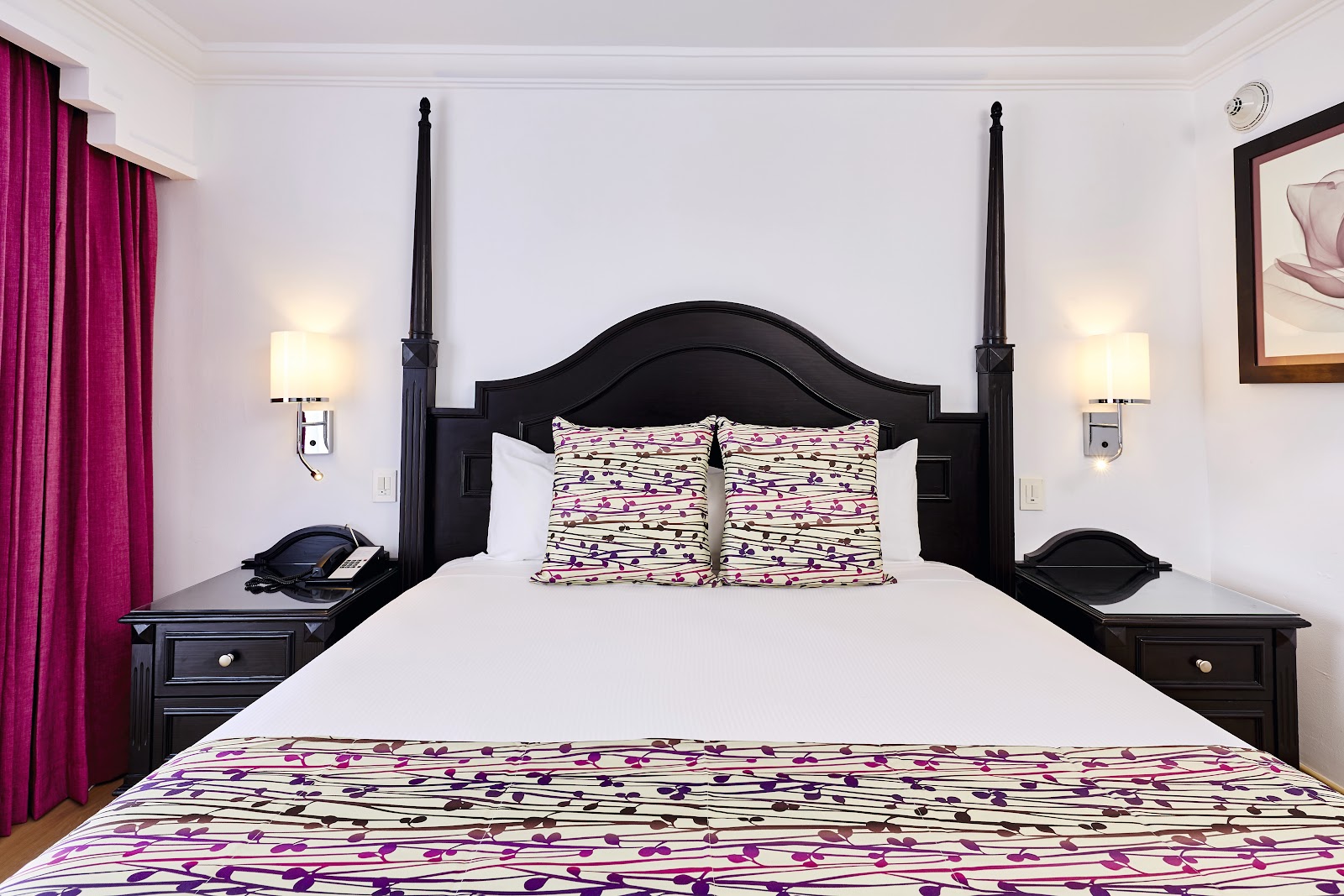 Black four-poster bed with patterned purple and white bedding in a resort guest room with magenta curtains and twin nightstands.