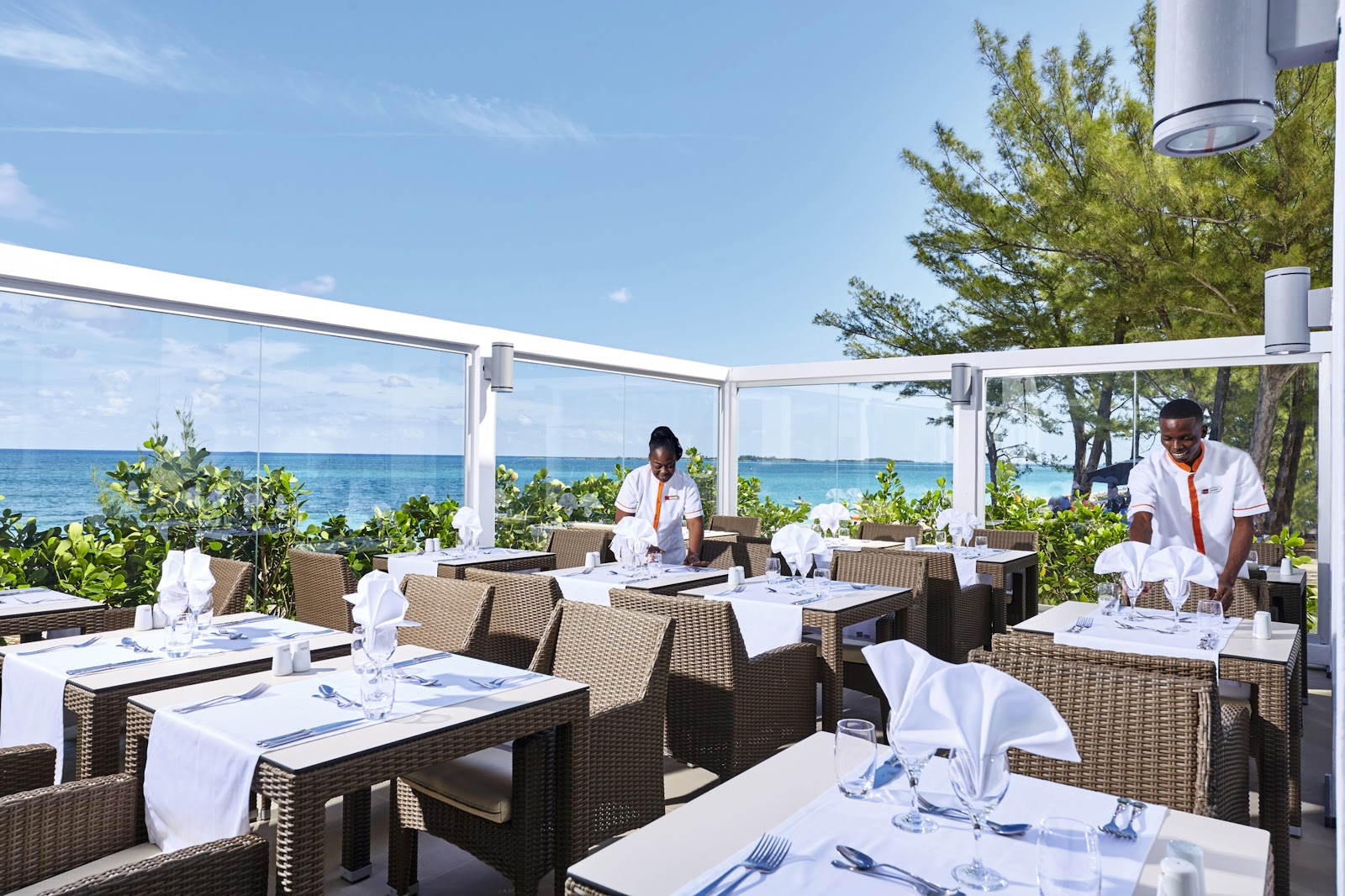 Beachfront dining area at Hotel Riu Palace Paradise Island with woven chairs, white linens, and turquoise ocean views beyond glass panels.