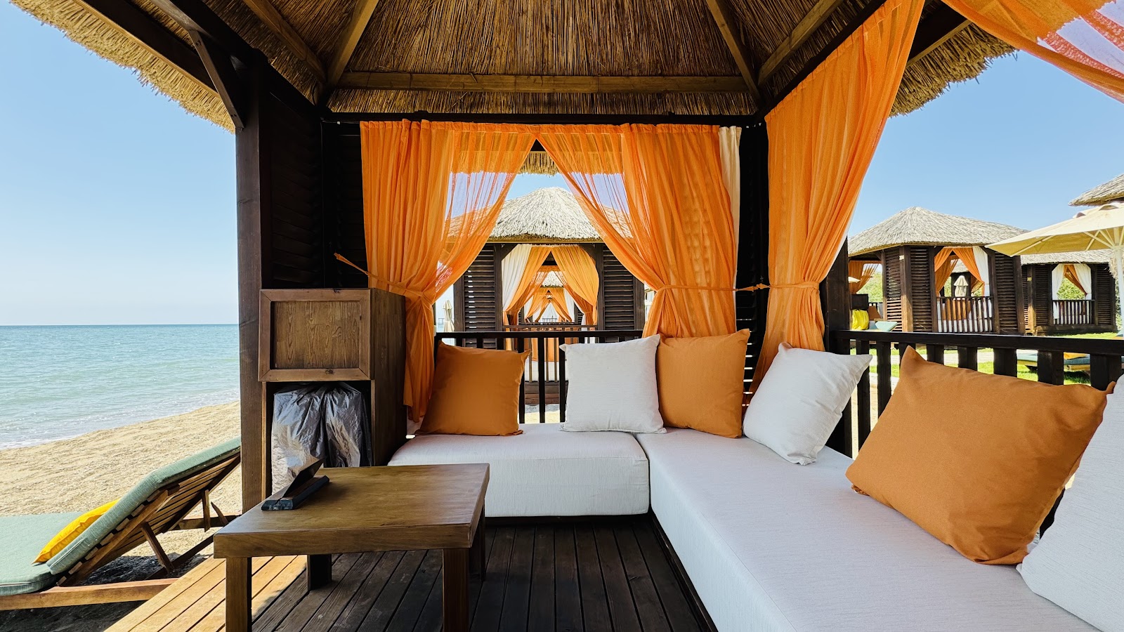 Beachfront cabana with orange curtains, white and orange cushioned seating, wooden table, and thatched roof overlooking the sea at Rixos Premium Belek.