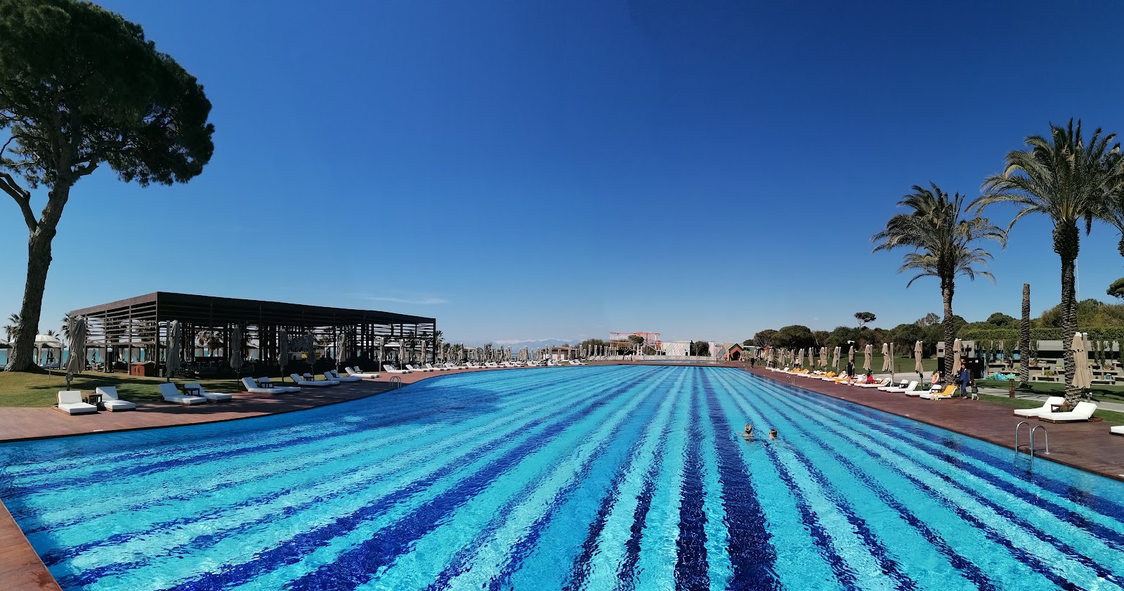 Striped pool deck at sunrise with palm trees, loungers, and a pavilion reflected in still water at Rixos Premium Belek.