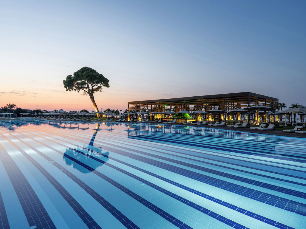 Large lap pool with lane dividers at twilight, a single pine tree, and the Rixos Premium Belek's lit pavilion visible across the water.