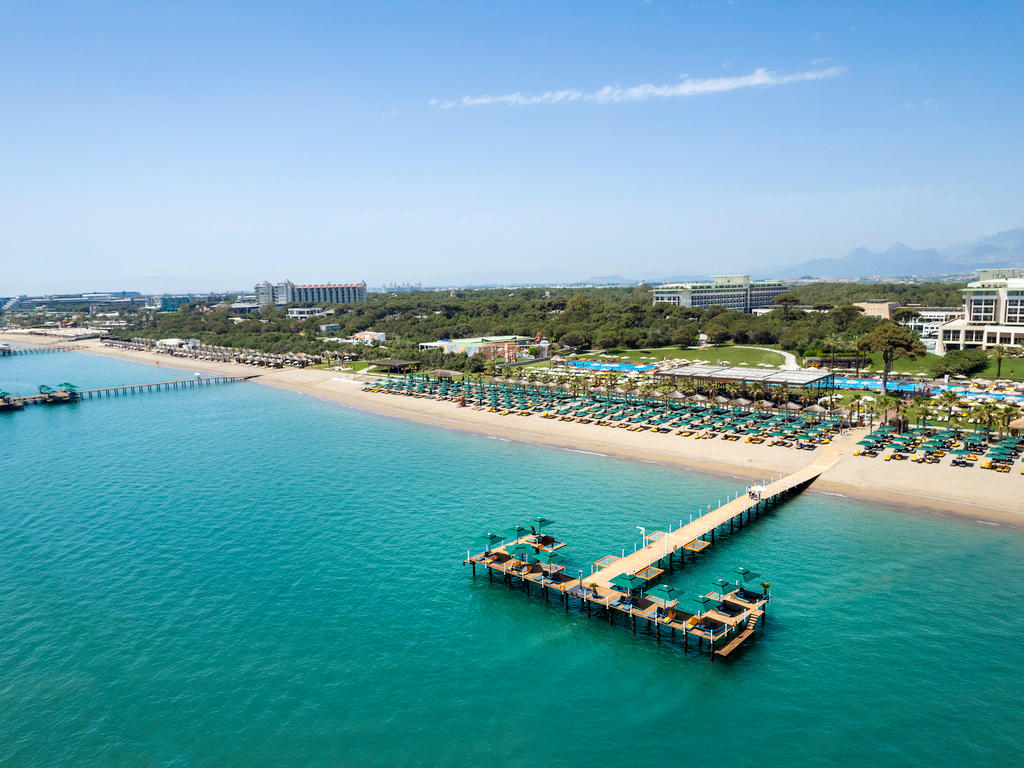 Sunset over multiple blue pools and tropical grounds at Rixos Premium Belek, with white hotel building and lush landscaping.