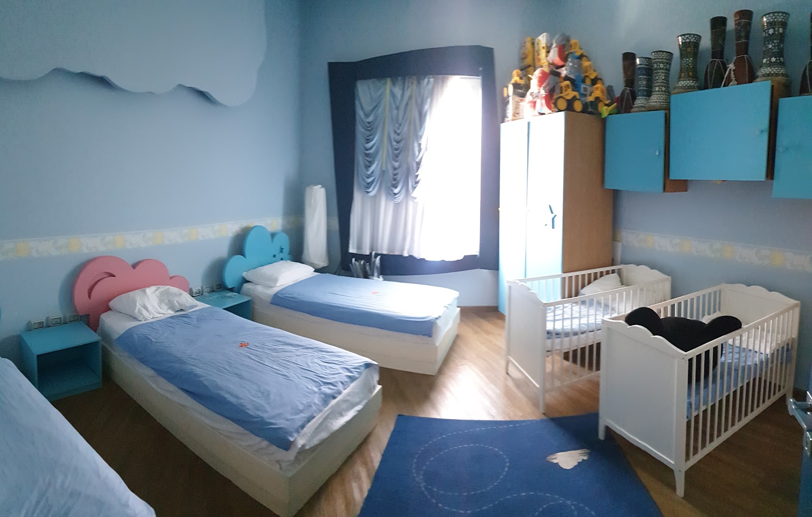 Light blue children's bedroom with two single beds, two white cribs, blue flooring, and cloud-shaped headboards at Rixos Premium Seagate.
