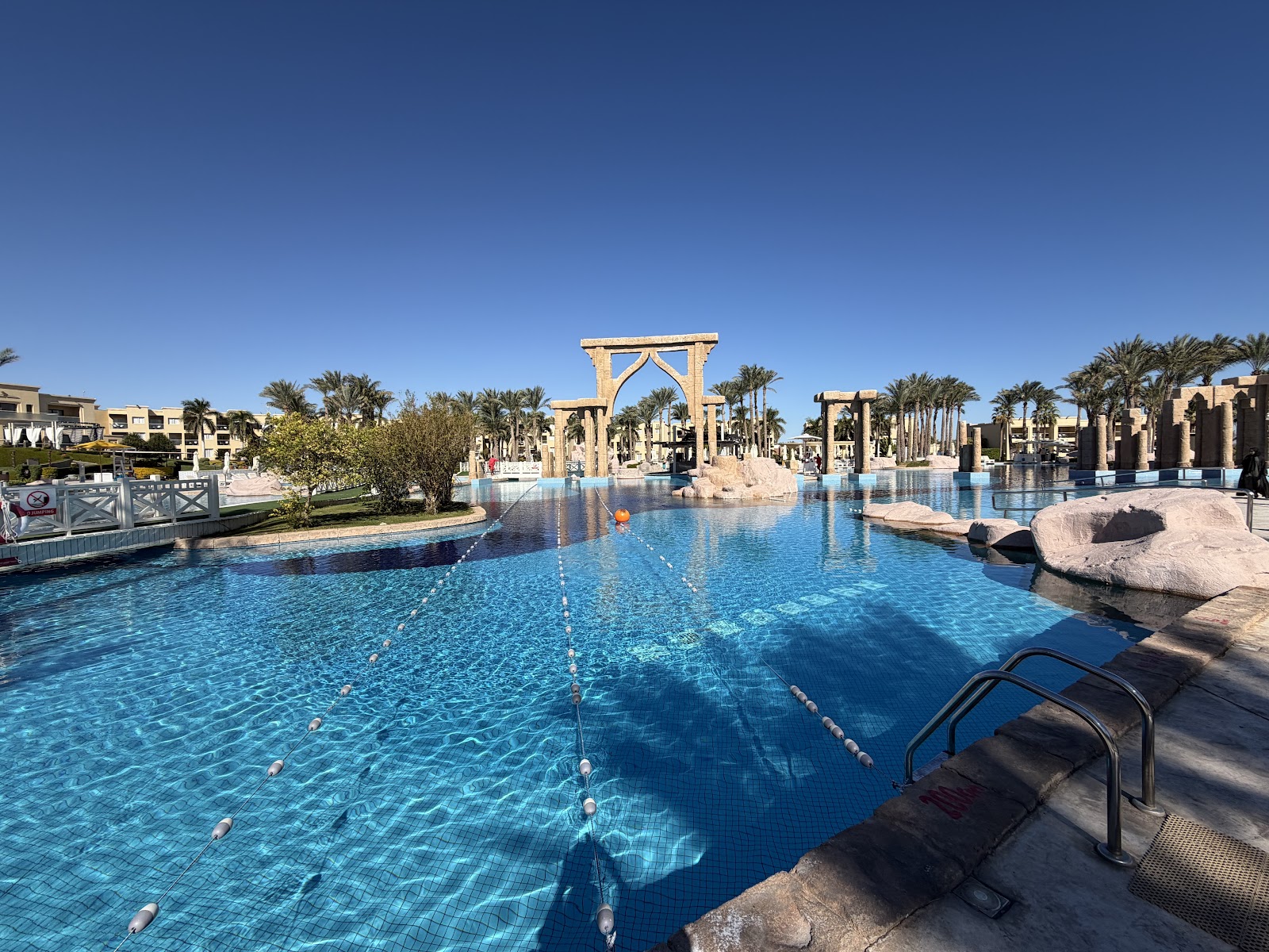 Large lagoon pool with Egyptian-themed stone archways and palm trees under blue sky at Rixos Premium Seagate.