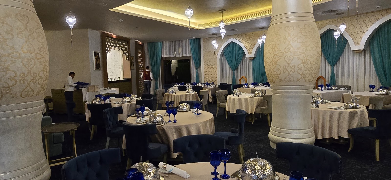Elegant banquet hall at Rixos Premium Seagate with round tables dressed in cream linens, blue accents, and ornamental centerpieces beneath a coffered ceiling with pendant lights.
