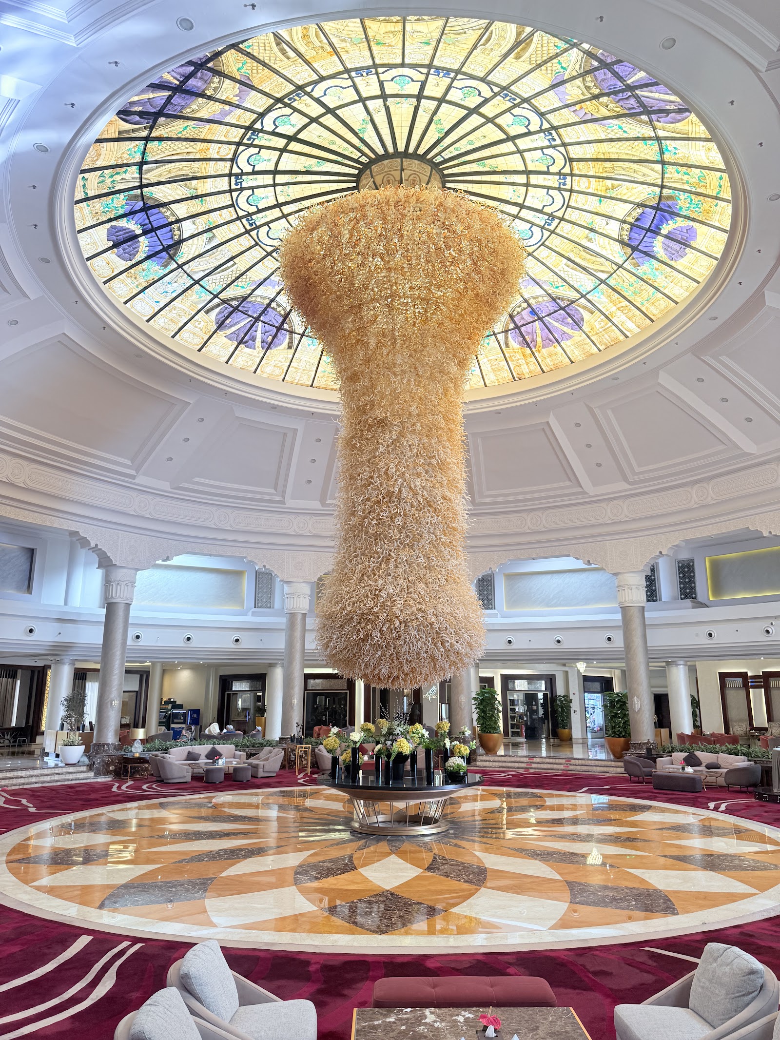 Grand lobby atrium with a massive woven pendant chandelier hanging from an ornate stained-glass dome, marble floors, and columns.