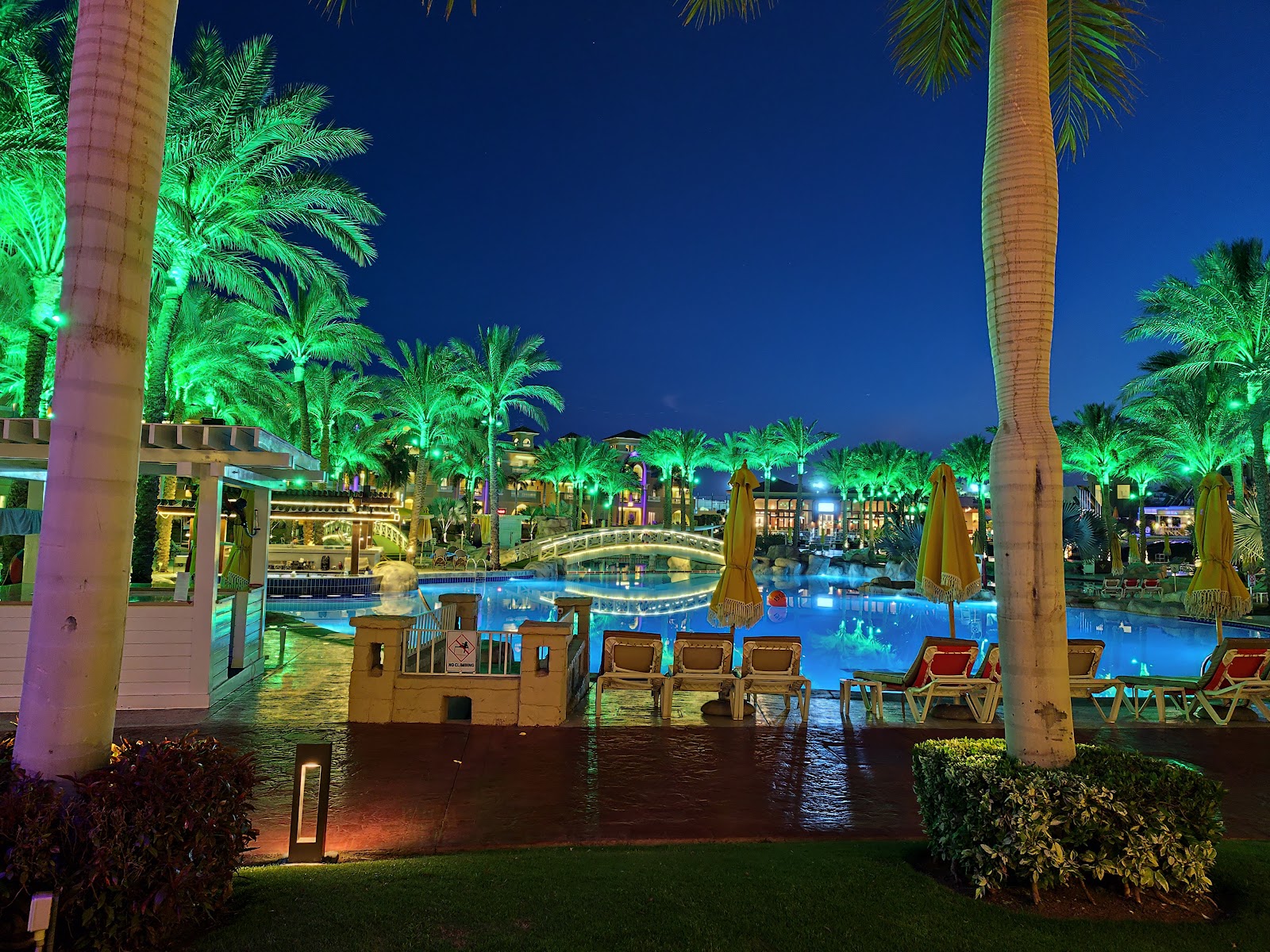Nighttime pool area at Rixos Premium Seagate with illuminated palms, lounge chairs, and a decorative castle structure reflected in the glowing blue water.