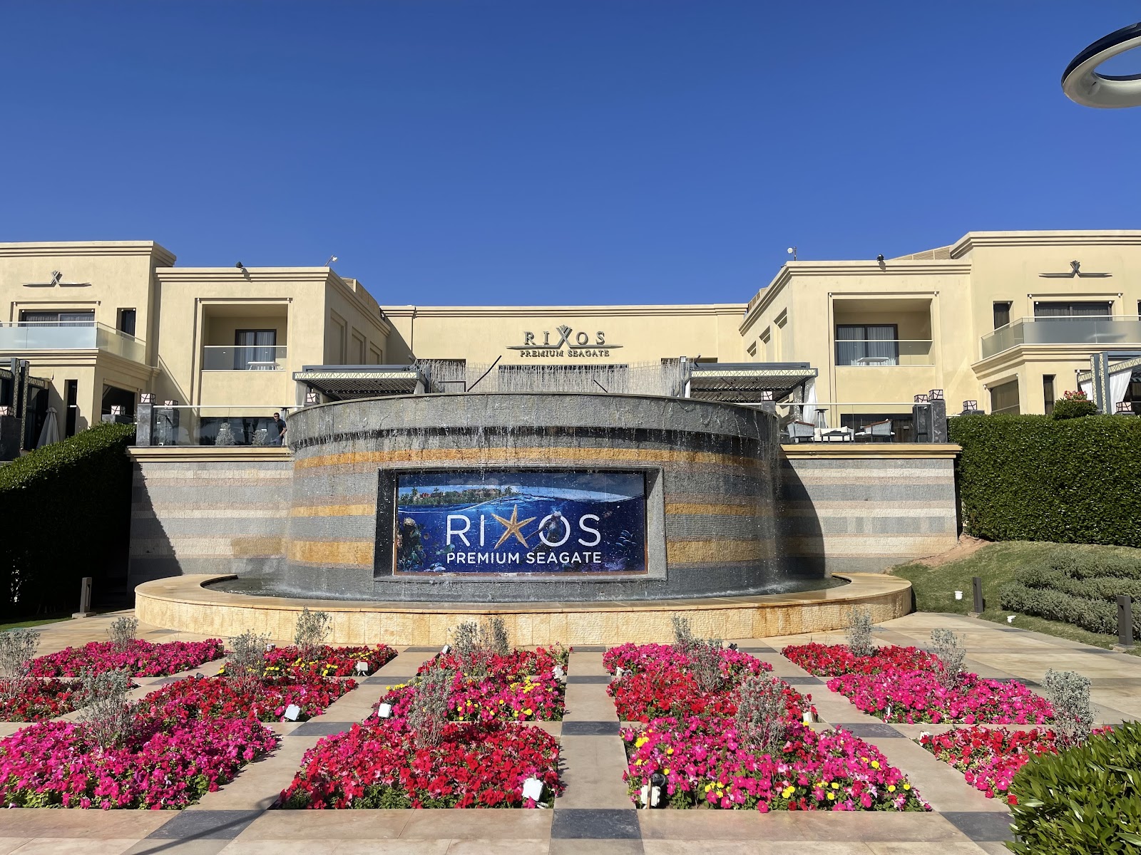 Rixos Premium Seagate entrance with stone signage, pink and red flowers in landscaped beds, and cream-colored resort buildings under clear blue sky.