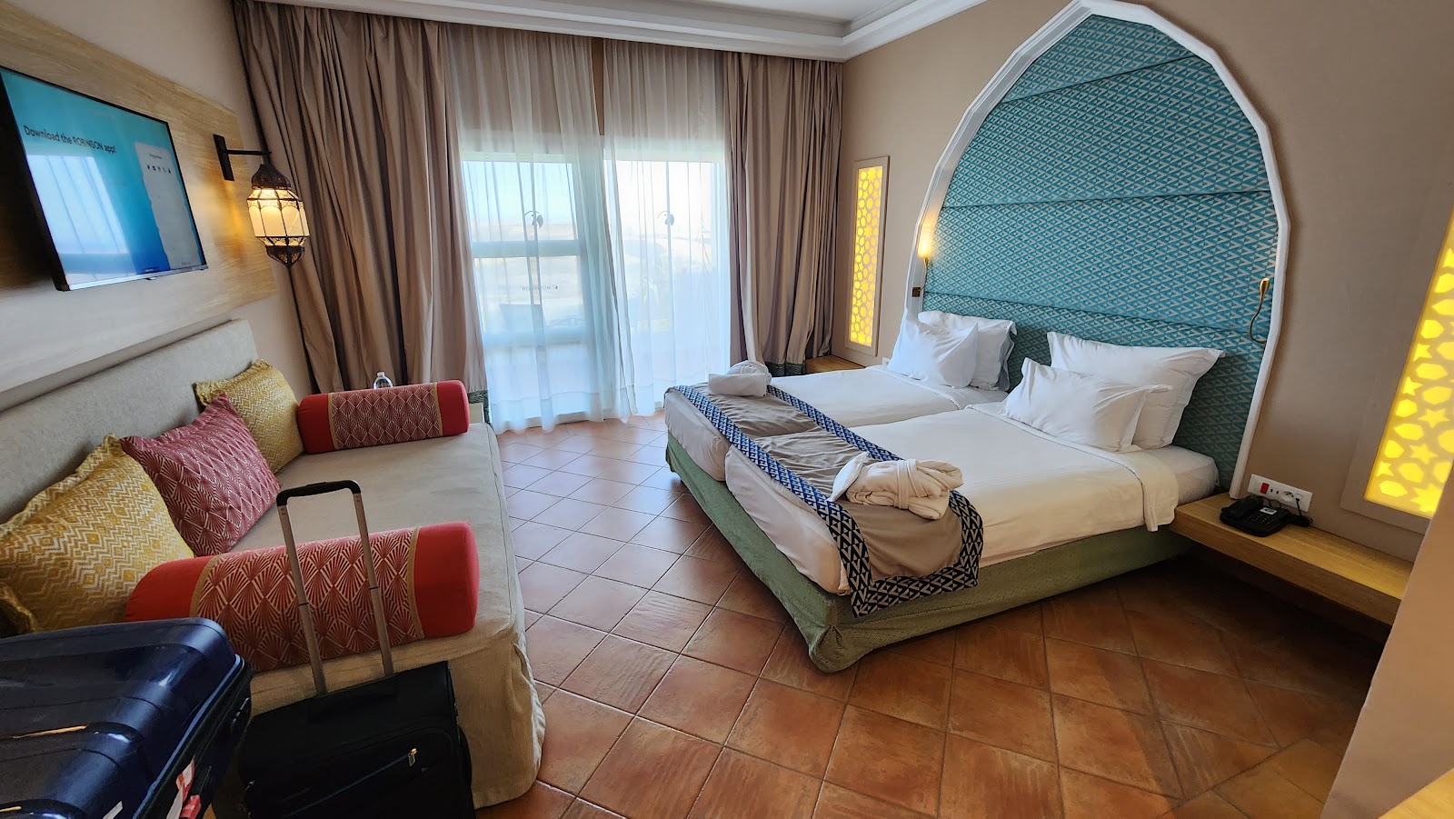 Resort bedroom with two double beds, teal headboard, sitting area with red and gold cushions, and balcony overlooking the ocean.