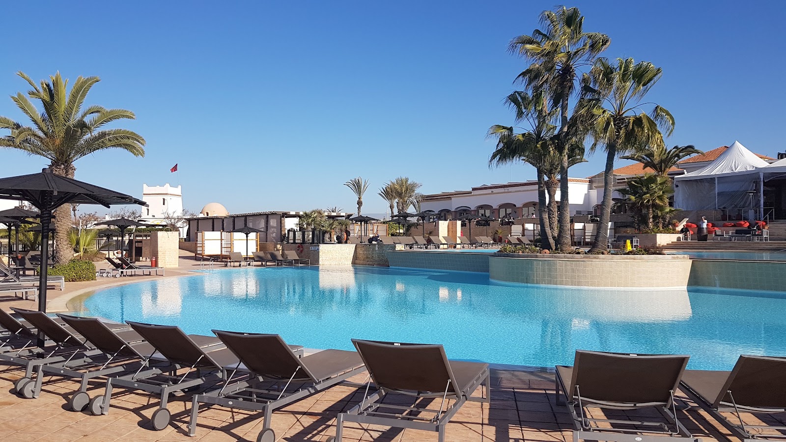 Lounge chairs line the pool deck at ROBINSON Agadir, with turquoise water, palm trees, and clay-colored resort buildings under clear blue sky.