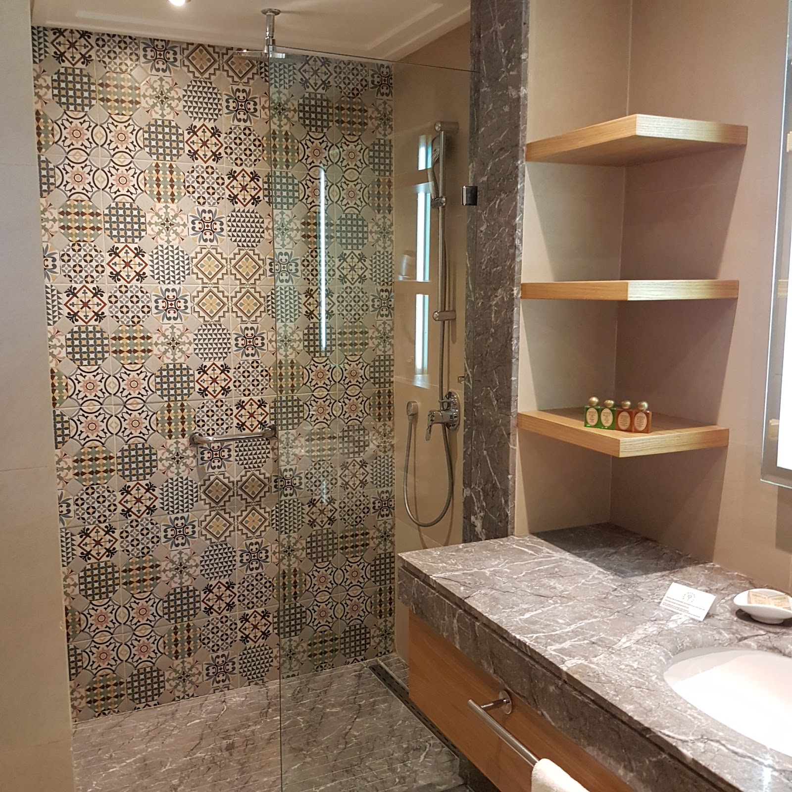 Modern bathroom with patterned tile walk-in shower, marble countertop, wooden shelving with toiletries, and sink at ROBINSON Agadir.