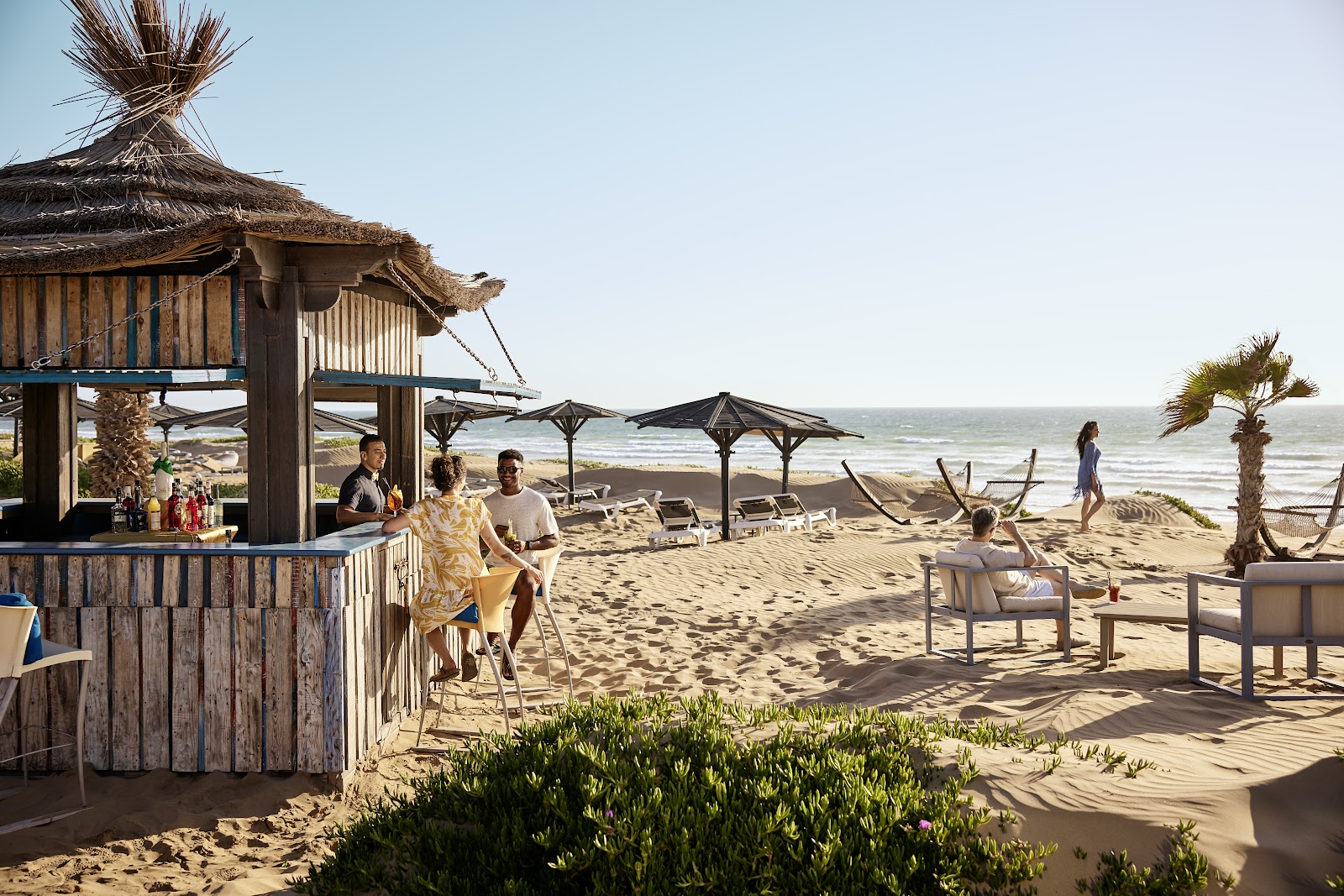 Beachfront bar with thatched roof at ROBINSON Agadir, guests relaxing under umbrellas on sand with ocean view.