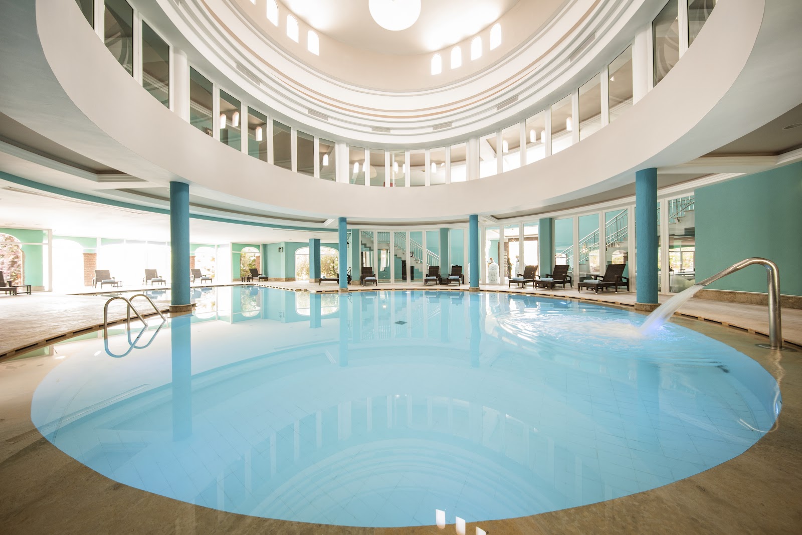 Indoor swimming pool with turquoise water, curved architecture, blue support columns, and loungers lining the sides under a domed skylight.