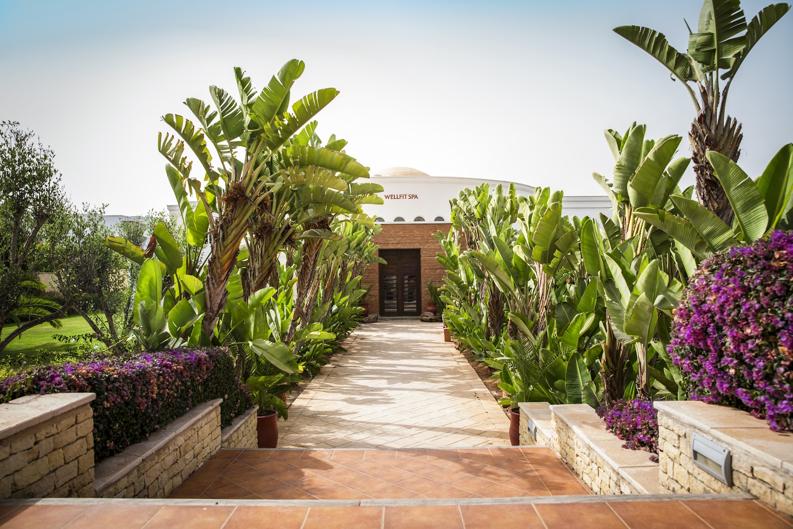 Shaded cabanas with white curtains line a lagoon at ROBINSON Agadir, surrounded by palm trees and tropical plants.