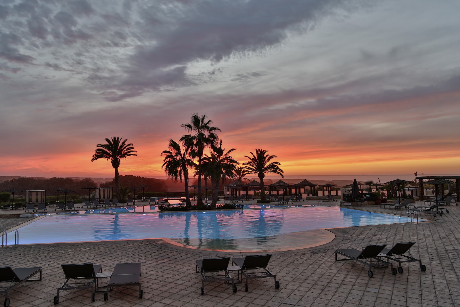 Resort pool with palm trees silhouetted against an orange and pink sunset sky, lounge chairs in foreground.