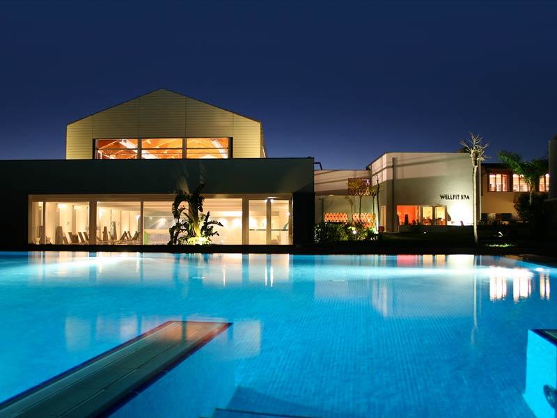 Illuminated modern buildings with floor-to-ceiling windows overlook a large glowing blue pool at dusk, with a diving board in the foreground.