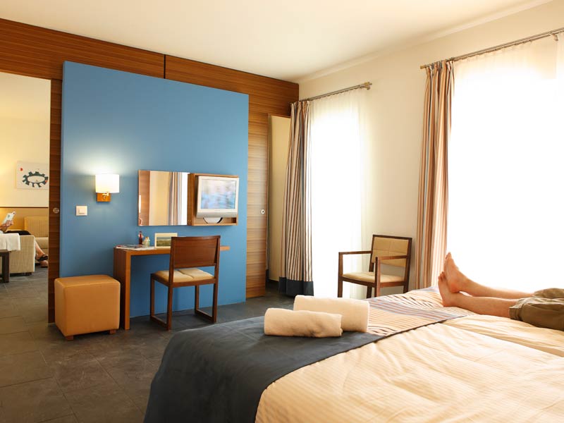 Resort bedroom with twin beds, blue accent wall, wooden desk area, orange ottoman, and bright windows with curtains.
