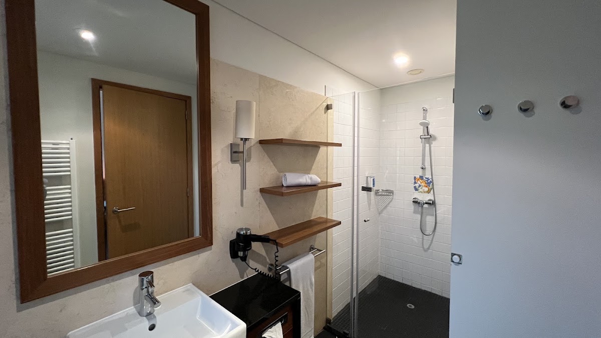 Modern bathroom with white sink, wooden-framed mirror, walk-in shower with white tile walls, and wooden shelving holding towels.