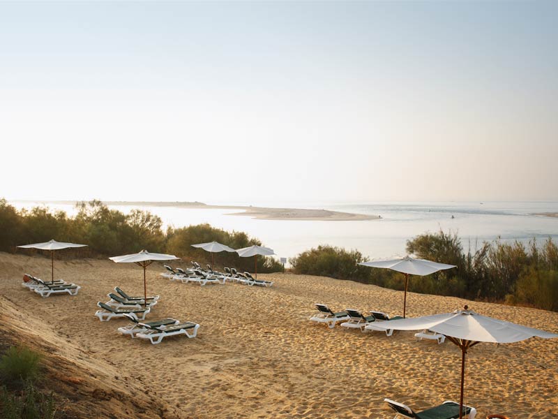 Sandy beach with white umbrellas and lounge chairs overlooking calm bay waters at Robinson Quinta da Ria.