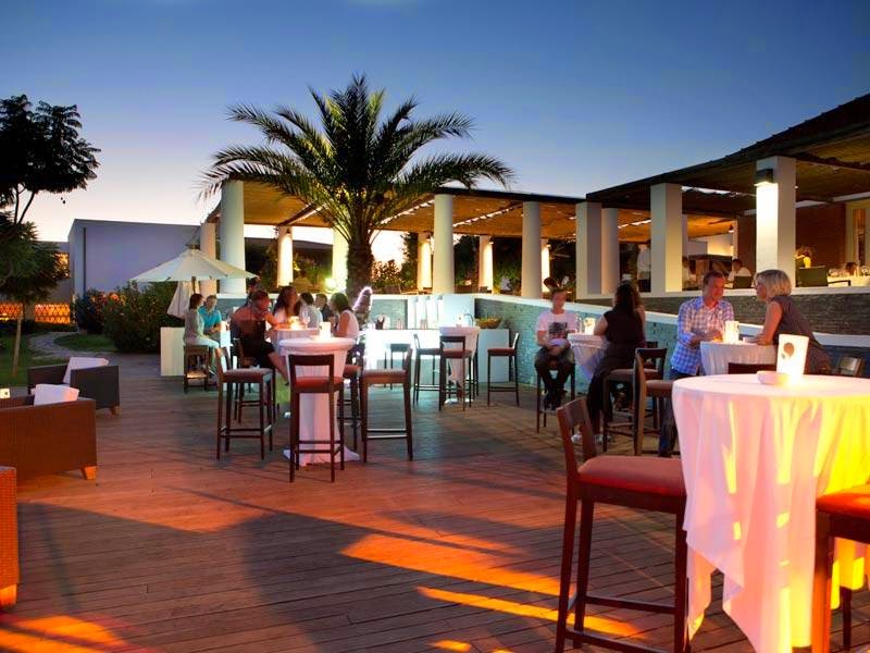 Guests gather on an illuminated terrace at dusk with a tall palm tree, bar seating, and glowing table accents under a covered pavilion.