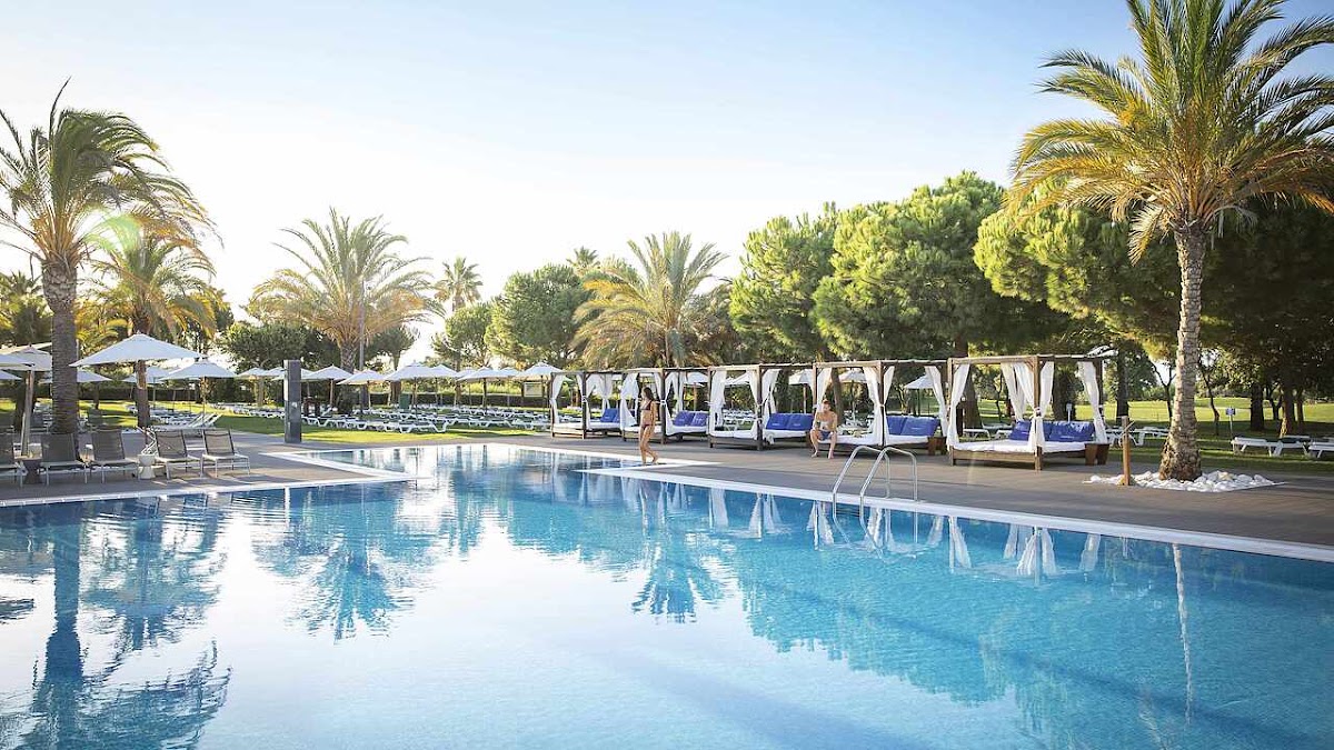 Resort swimming pool with blue cabanas, white umbrellas, and palm trees under clear sky.