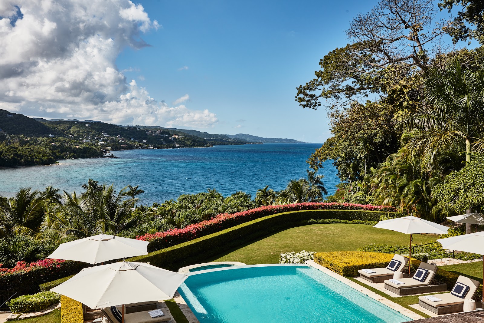 Turquoise pool with white umbrellas overlooks manicured gardens and blue Caribbean Sea at Round Hill Hotel and Villas.