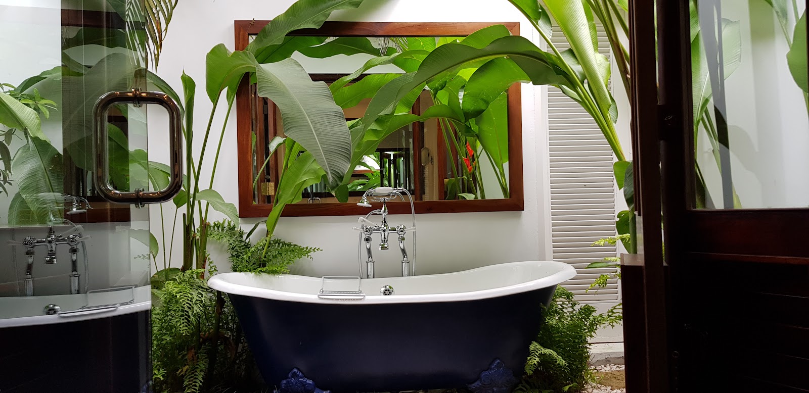 Dark freestanding bathtub with chrome faucet in a bathroom surrounded by lush green plants and banana leaves at Round Hill Hotel and Villas.