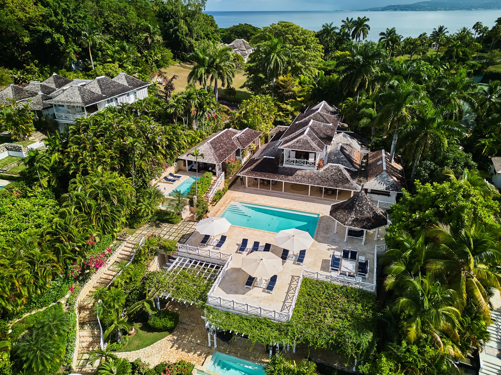Round Hill Hotel and Villas villa with peaked roof and white shutters overlooks turquoise Caribbean waters, surrounded by palm trees and tropical landscaping.