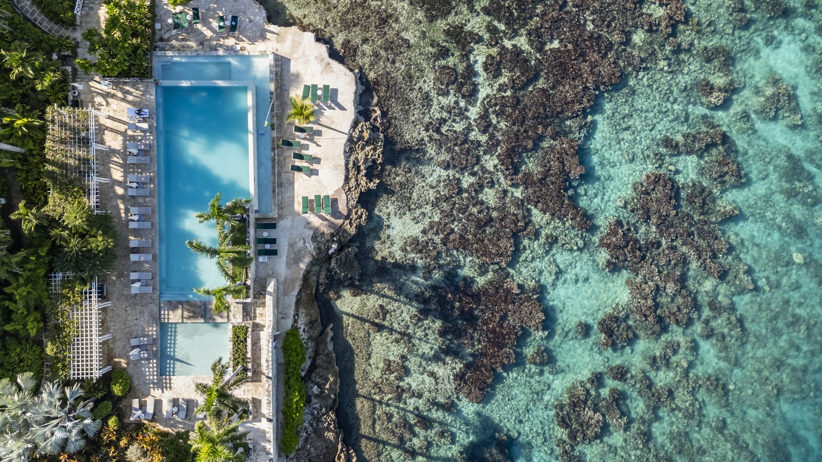 Aerial view of Round Hill Hotel and Villas nestled on a tropical coastline with white buildings, pool, and beach surrounded by dense green vegetation and mountains.