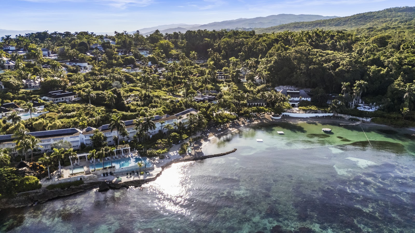 Aerial view of Round Hill Hotel and Villas showing stone-roofed buildings, turquoise pools, lounge chairs, lush tropical vegetation, and the Caribbean Sea beyond.
