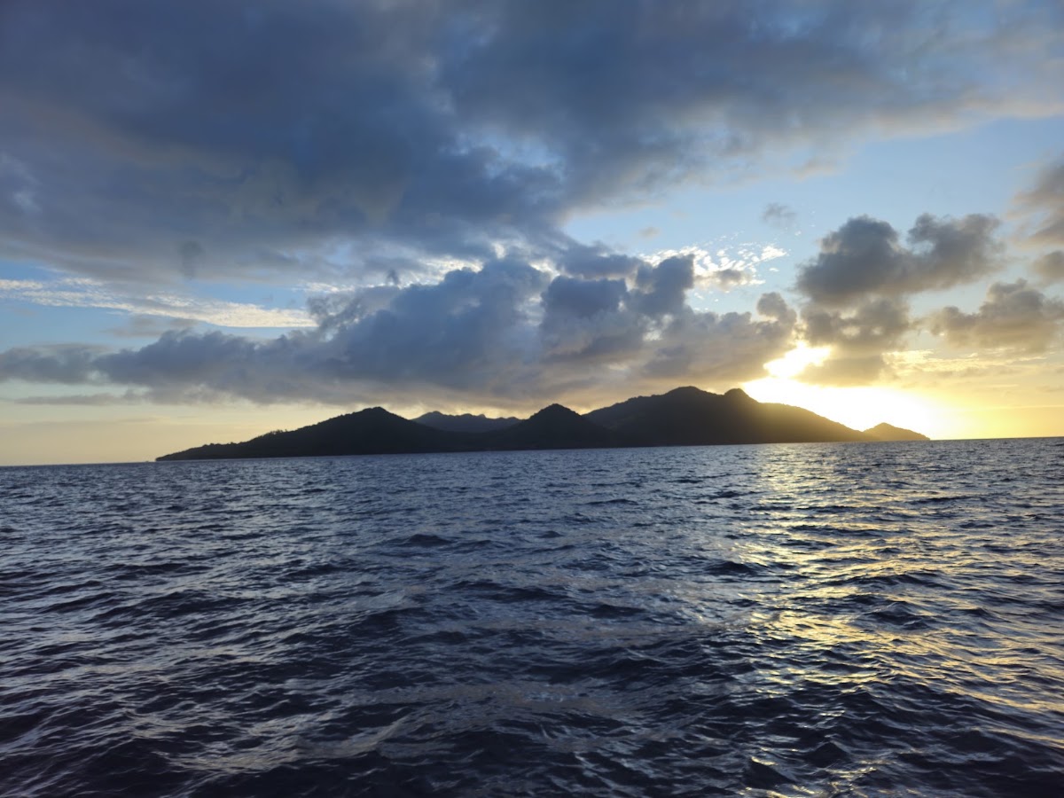 Sunset view across calm ocean waters toward a dark mountainous island silhouetted against golden and cloudy sky.