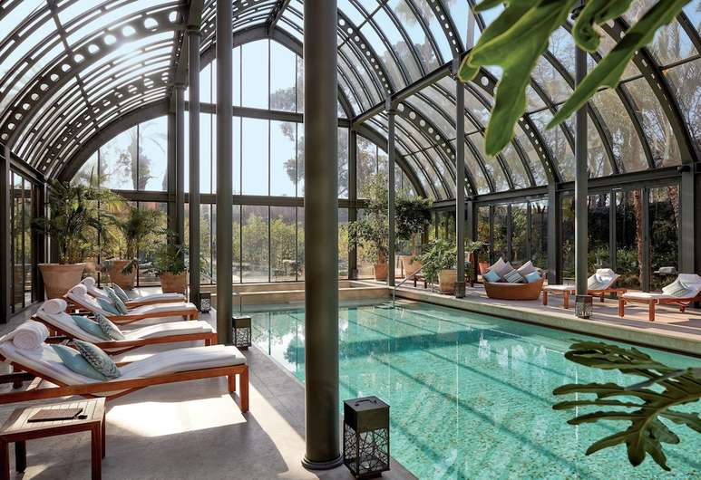 Indoor pool at Royal Mansour Marrakech with wooden loungers, arched glass ceiling, and views of gardens and mountains beyond.