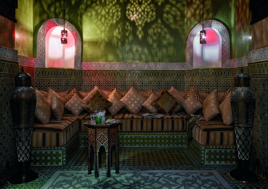 Moroccan-style lounge with green zellige tilework, arched alcoves with pink lighting, brown cushioned seating, ornate black lanterns, and decorative table.