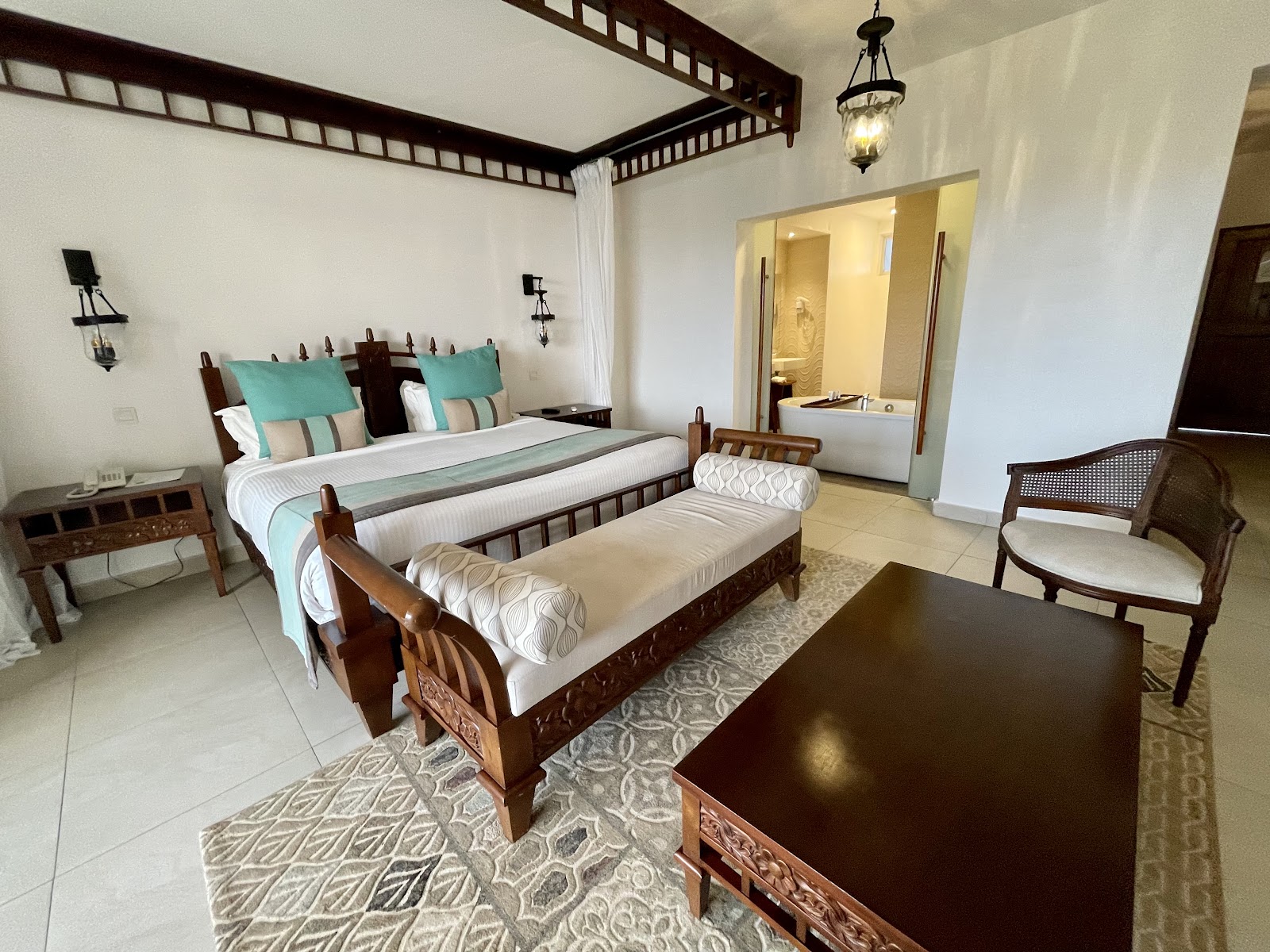 Resort suite with four-poster bed draped in turquoise linens, bench seating, dark wood furniture, and en-suite bathroom visible through open doors.