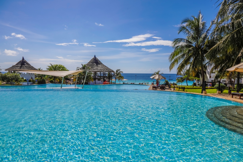 Resort pool with thatched-roof structures, palm trees, and clear ocean view beyond at Royal Zanzibar Beach Resort.