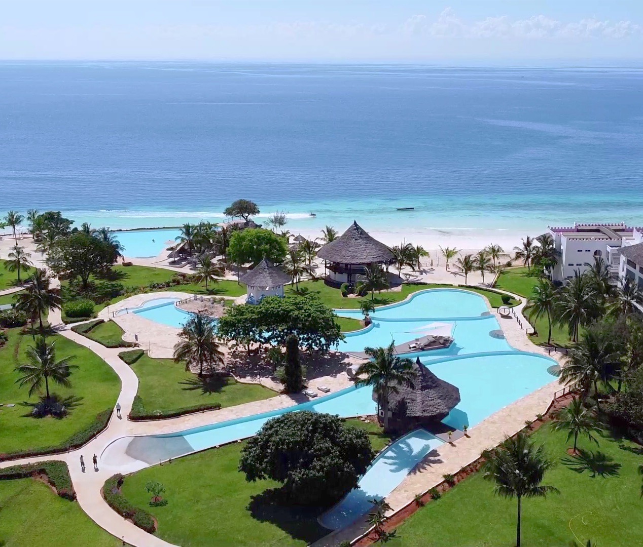 Aerial view of Royal Zanzibar Beach Resort showing turquoise pools, manicured lawns, palm trees, thatched-roof structures, and white sandy beach with Indian Ocean beyond.