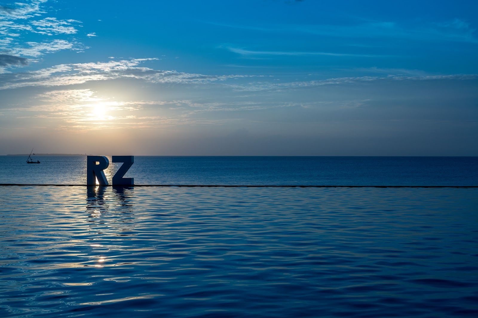 RZ letters stand in shallow water at sunset with a sailboat and calm ocean under a blue sky.