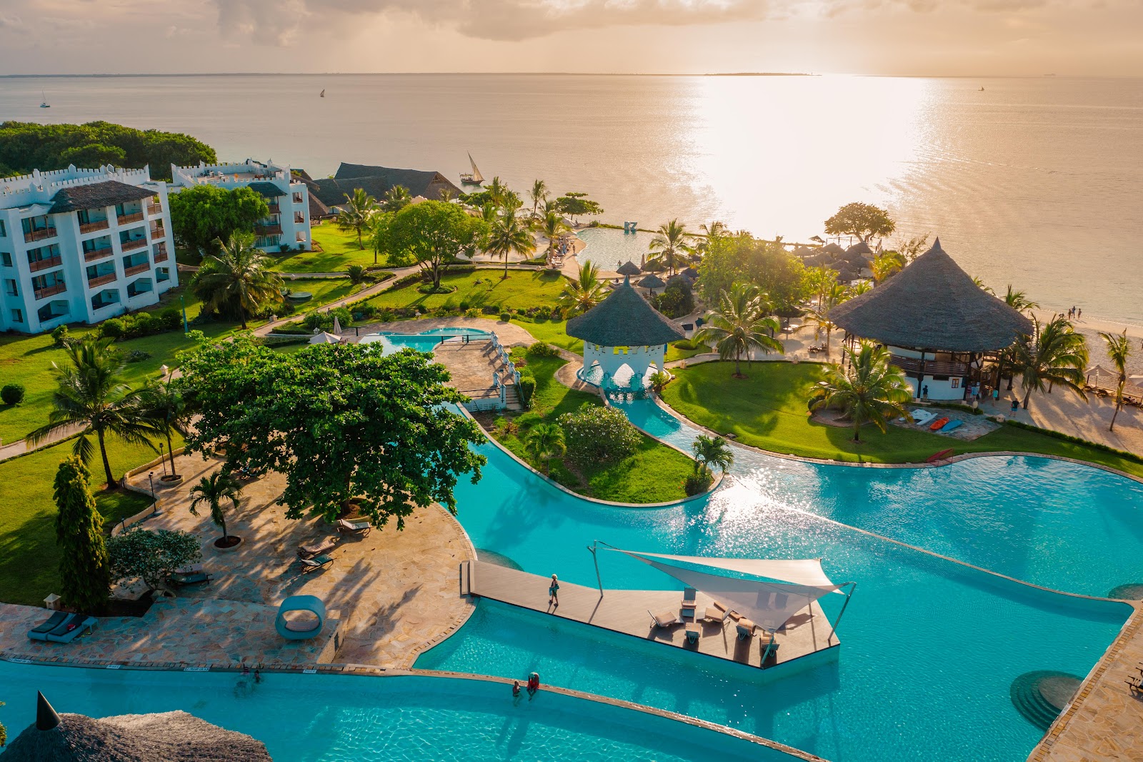 Aerial view of Royal Zanzibar Beach Resort with turquoise lagoon pools, thatched-roof structures, white beachfront buildings, and Indian Ocean beyond at sunset.