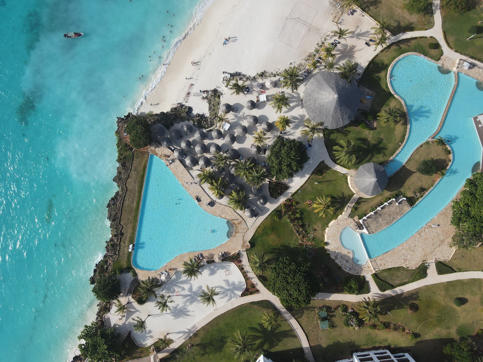 Aerial view of Royal Zanzibar Beach Resort showing turquoise ocean, white sand beach, multiple pools, palm trees, and thatched-roof structures.