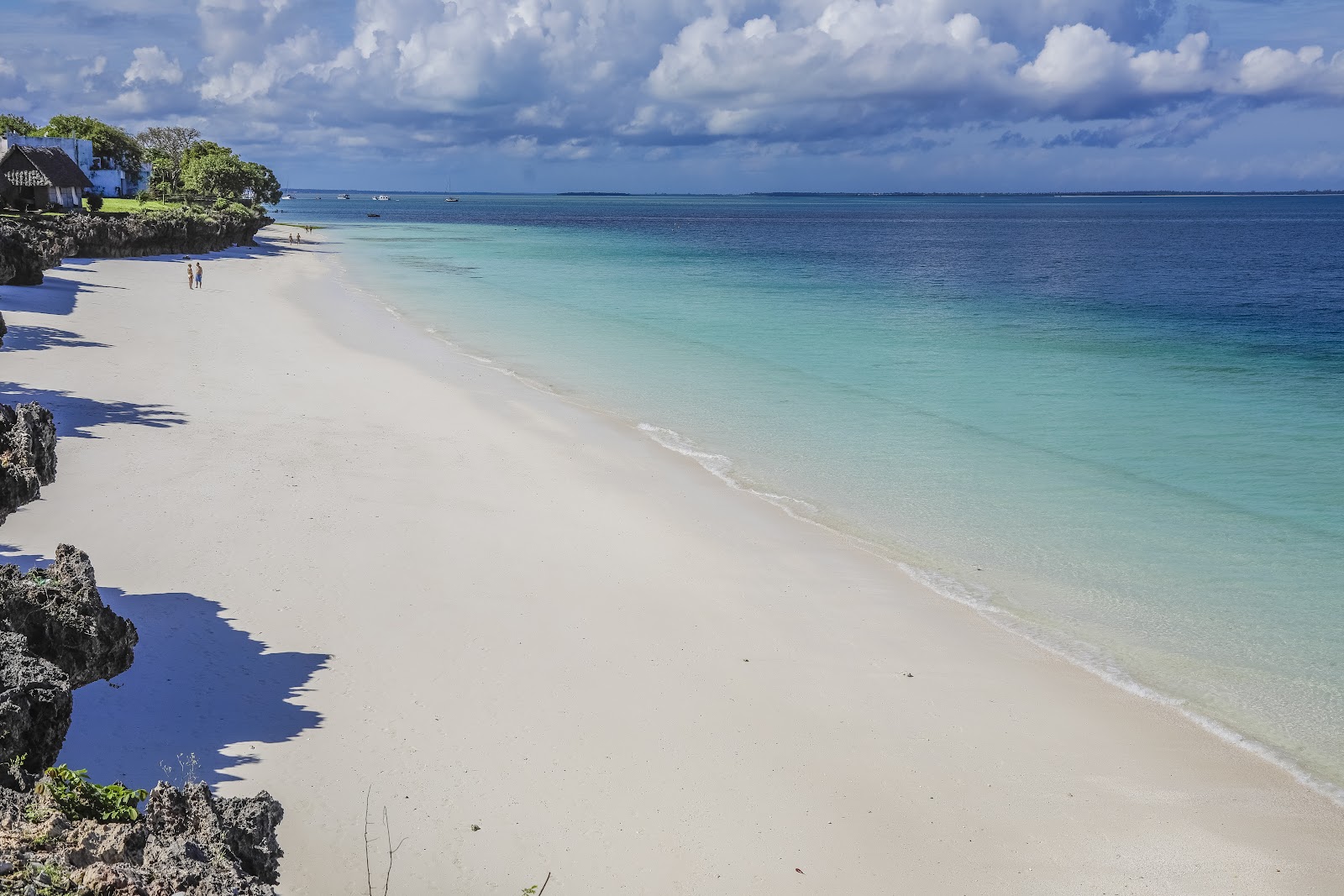 White sand beach with turquoise water and dark rock formations, a few guests wading, resort building visible among trees on the left.