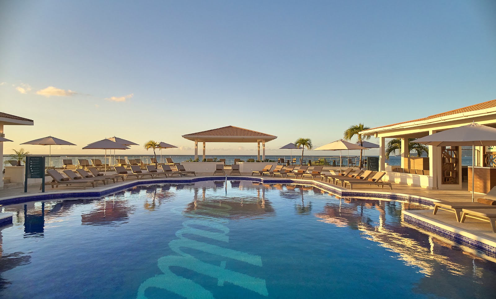 Royalton Grenada pool deck at sunset with loungers, umbrellas, gazebo, and ocean view beyond.