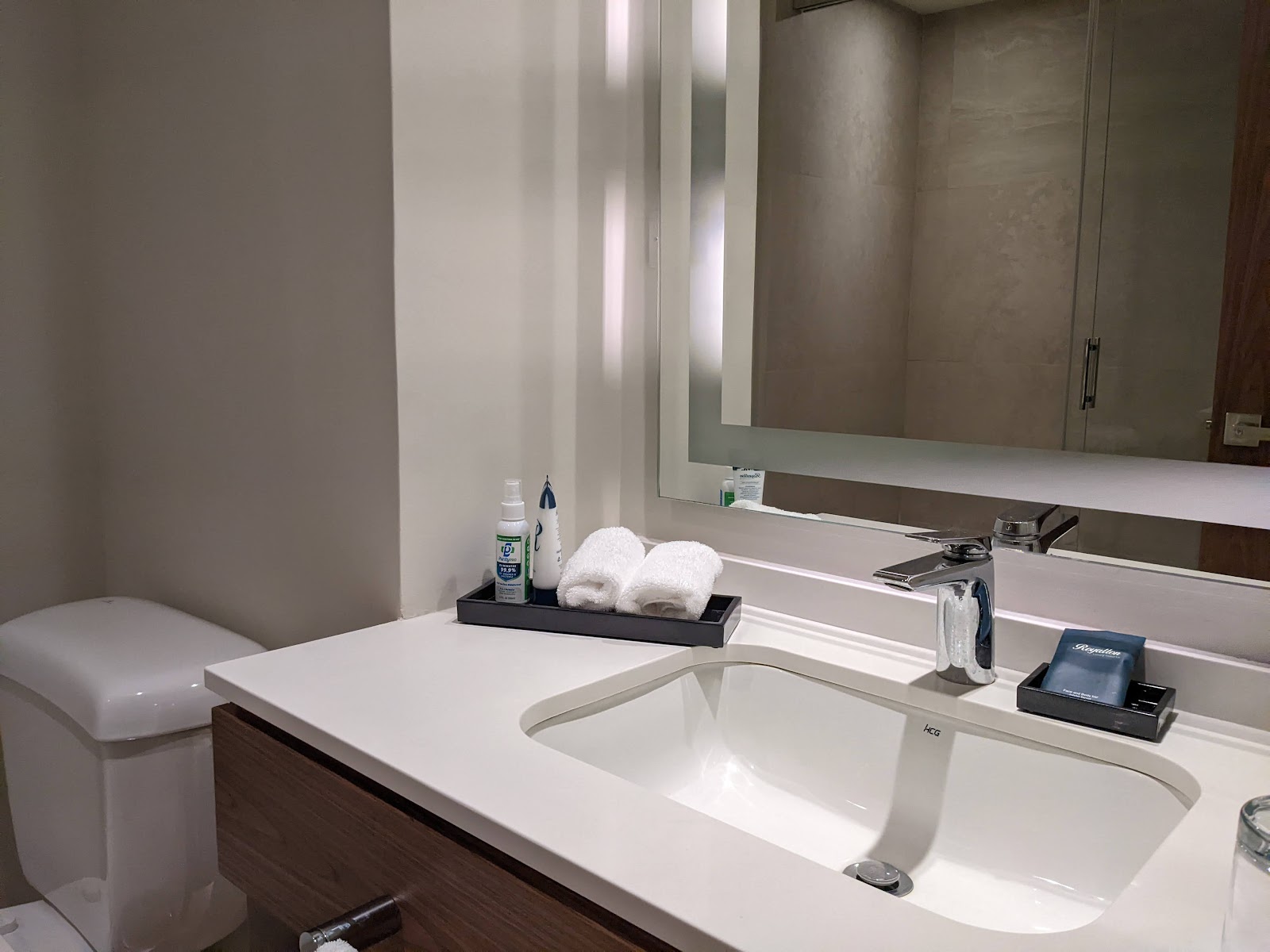 Modern bathroom at Royalton Grenada with white sink, dark wood vanity, large mirror, folded towels, and toiletries on black tray.