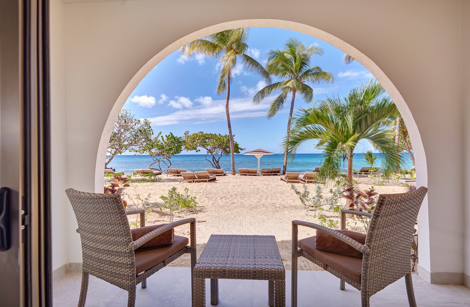 Woven lounge chairs and table on a beachfront patio at Royalton Grenada, with palm trees and ocean view beyond.