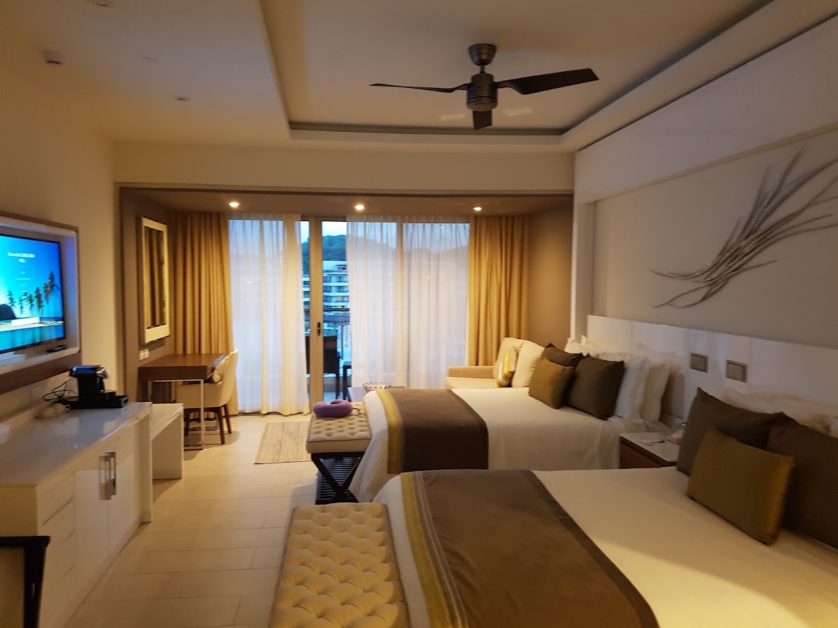 Resort guest room with two beds, yellow and brown pillows, balcony doors with sheer curtains, ceiling fan, and ocean view at Royalton Saint Lucia.