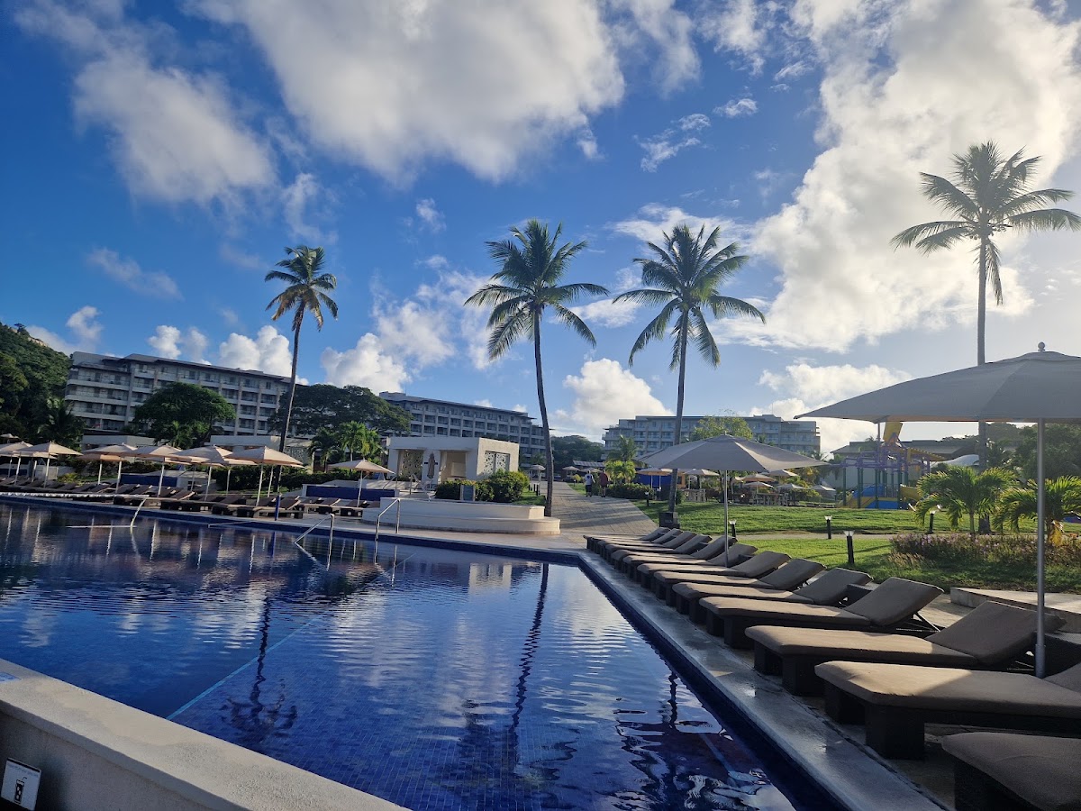 Royalton Saint Lucia pool with blue water, black lounge chairs, palm trees, and multi-story resort buildings under a blue sky with white clouds.