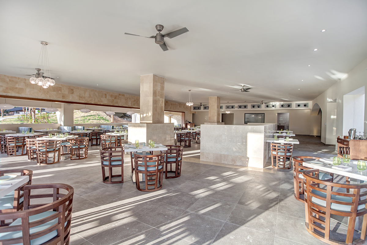 Modern dining room at Royalton Saint Lucia with wooden chairs at round tables, concrete columns, and natural light streaming through large windows.