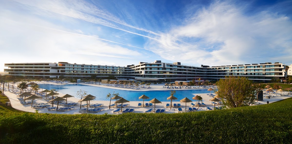 Modern curved resort building overlooking a large lagoon-style pool with thatched umbrellas, lounge chairs, and manicured grounds under blue sky.